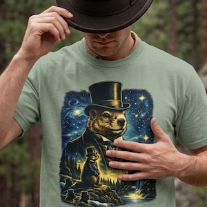 May include: A sage green t-shirt featuring a graphic print of a groundhog wearing a black top hat and suit. The design includes a starry night sky background, a smaller groundhog figure in a forest landscape, and a flowing stream at the bottom.