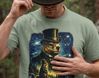 Puede incluir: Una camiseta verde salvia con un estampado gráfico de una marmota con sombrero de copa y traje negro. El diseño incluye un cielo nocturno estrellado, una marmota más pequeña en un paisaje forestal y un arroyo que fluye en la parte inferior.
