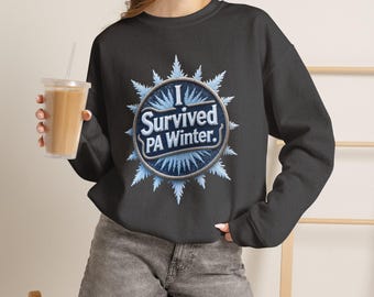 Puede incluir: Una sudadera negra de manga larga con cuello redondo que presenta un gráfico circular central con un borde de copos de nieve azul claro. El texto dentro del círculo dice I Survived PA Winter en letras blancas sobre fondo azul.