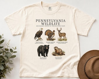 Puede incluir: Una camiseta de color crema con un estampado gráfico titulado PENNSYLVANIA WILDLIFE RECOGNIZED SPECIES OF THE KEYSTONE STATE. El diseño muestra ilustraciones de un águila calva, un urogallo, un ciervo de cola blanca, un oso negro y una marmota.