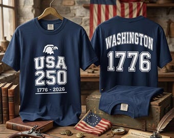 Puede incluir: Una camiseta azul marino con un gráfico blanco de una cabeza de águila sobre el texto USA 250 y 1776-2026. La parte posterior de la camiseta muestra la palabra WASHINGTON en letras blancas arqueadas sobre los grandes números blancos 1776.
