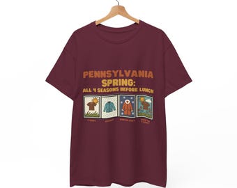 Puede incluir: Una camiseta granate con el texto "PENNSYLVANIA SPRING: ALL 4 SEASONS BEFORE LUNCH" en naranja. Debajo del texto hay cuatro pequeñas imágenes que representan las estaciones. La camiseta está colgada en una percha de madera.