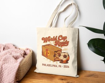 Puede incluir: Bolso tote color crema con un diseño retro. Presenta las palabras "World Cup Ready" en escritura naranja, una caja de pretzels y un balón de fútbol. El texto "PHILADELPHIA, PA - 2026" está impreso en la parte inferior.