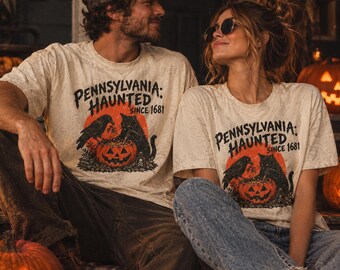 Puede incluir: Dos camisetas de manga corta color hueso con un estampado gráfico que incluye el texto "PENNSYLVANIA: HAUNTED SINCE 1681" sobre una ilustración de un gato negro y un cuervo junto a una calabaza naranja brillante con forma de linterna.
