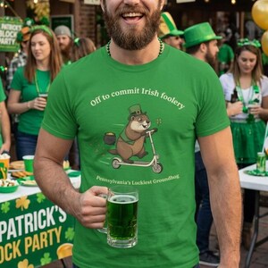 May include: Green t-shirt with the text "Off to commit Irish foolery" and a cartoon groundhog riding a scooter. The shirt also says "Pennsylvania's Luckiest Groundhog." The person is holding a glass of green beer.