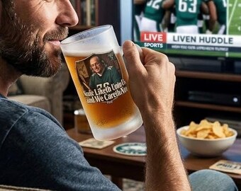 Puede incluir: Una jarra de cerveza de vidrio esmerilado con asa, llena de cerveza dorada. La jarra presenta un gráfico de una persona con una camiseta verde y el texto: "None Liketh Unto Us and We Careth Not". El vidrio tiene un acabado texturizado y semitransparente.