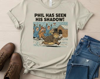 Puede incluir: Camiseta beige con una ilustración de dibujos animados de una marmota con sombrero de copa huyendo de una multitud. El texto "PHIL HAS SEEN HIS SHADOW!" está impreso encima de la ilustración. La camiseta se muestra con vaqueros azules y botas de cuero marrones.