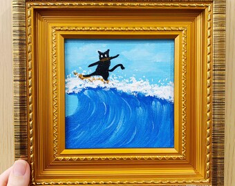 May include: A framed painting depicts a black cat surfing a blue wave under a light blue sky. The cat is on a surfboard. The painting is in a gold-colored ornate frame. The artwork is square.