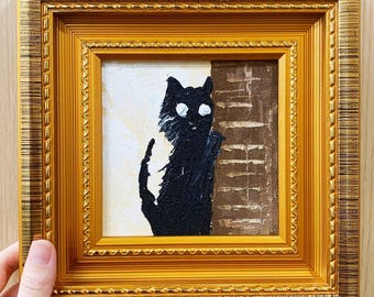 May include: A framed painting of a black cat with white eyes and a white chest, set against a beige and brown background. The artwork is in a gold-colored ornate frame, held by a hand.