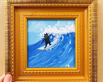 May include: A framed painting depicts a black cat surfing a blue wave. The cat is on a white surfboard, with a gold frame. The artwork is a square, with a blue sky background.