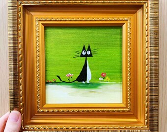 May include: A framed painting featuring a whimsical black and white cat with large eyes against a bright green background. The artwork is set in a gold ornate frame, with small flowers at the base of the painting. The overall style is cartoonish and charming.