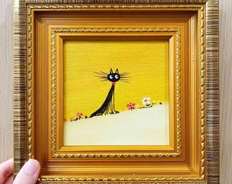 May include: A framed painting featuring a whimsical black cat with a red collar against a yellow backdrop. The cat is depicted with long whiskers and a slender body, with small flowers at the base of the hill. The frame is gold.