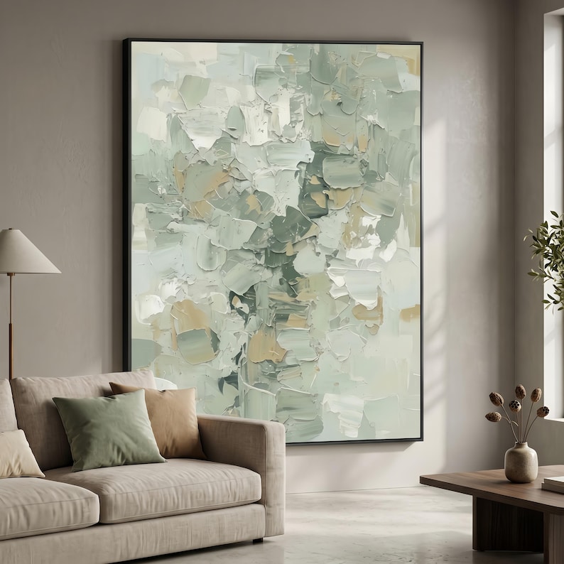May include: Large framed abstract painting with a textured design in shades of green, white, and tan. The artwork is displayed in a living room setting, with a sofa and cushions in the foreground. The painting is in a black frame.