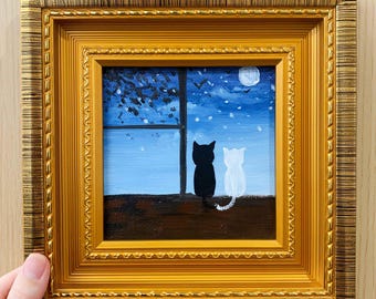 May include: A framed painting depicts two cats, one black and one white, silhouetted against a night sky with a full moon. The cats are perched on a windowsill, gazing out at the scene. The frame is a decorative gold color.