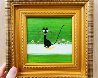 May include: A framed painting featuring a whimsical black cat with a pink collar, set against a green background with flowers. The cat has large eyes and a long tail. The frame is gold-colored with ornate detailing.