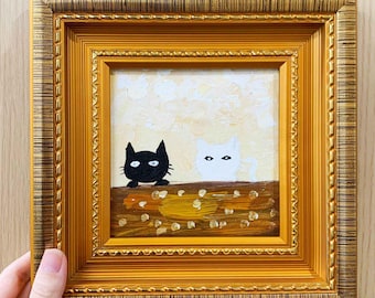 May include: Square framed painting featuring two cartoon cats, one black and one white, peeking over a brown ledge. The background is a light yellow with white brushstrokes. The frame is gold with ornate detailing.