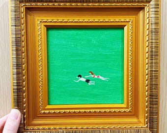 May include: A framed painting featuring two figures swimming in a green body of water. The figures are depicted in a minimalist style, with one wearing black shorts and the other a red and white bikini. The painting is set within a gold-colored ornate frame.