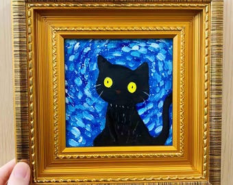 May include: A framed painting of a black cat with yellow eyes against a blue and white background. The cat is centered in the painting, and the frame is gold-colored with a detailed pattern.