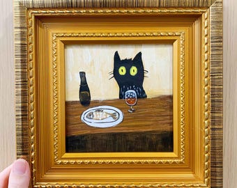 May include: A framed painting depicts a black cat with yellow eyes at a table with a bottle, a plate with a fish, and a glass of wine. The painting is in a gold-colored ornate frame.