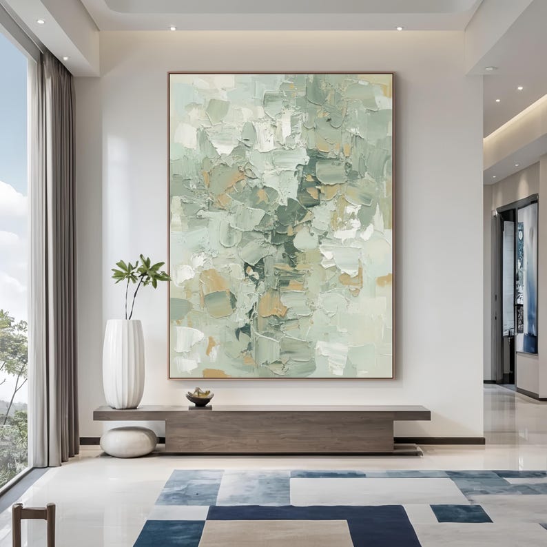 May include: Large abstract painting with a textured surface, in shades of green, beige, and white. The artwork is displayed in a room with a modern aesthetic, complemented by a wooden console and a patterned rug.
