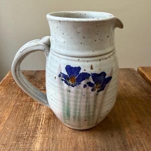 May include: A speckled, off-white ceramic jug with a handle and spout. The jug is decorated with blue floral designs and green vertical lines. The jug is sitting on a wooden surface.