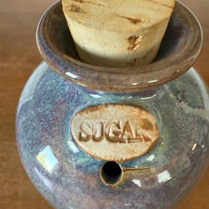 May include: A small, heart-shaped ceramic sugar bowl with a cork stopper. The bowl is glazed in shades of blue and purple with a brown rim. The word "SUGAR" is embossed on the front.