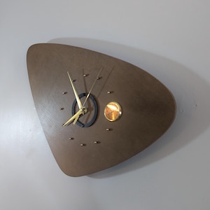 May include: A triangular wall clock with a smooth brown wooden face. It features gold-colored hands, small gold-colored dot hour markers, a black circular accent ring around the center, and a small circular gold-colored dial on the right side.