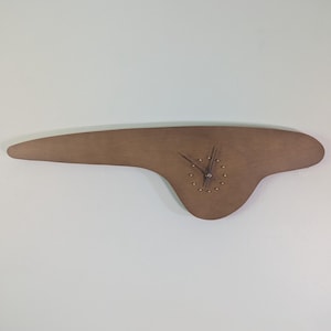 May include: A wall clock with an asymmetrical, elongated wooden body in a medium brown tone. The clock face features small, circular metallic markers for the hours and two thin, dark-colored hands positioned in the center of the wooden form.