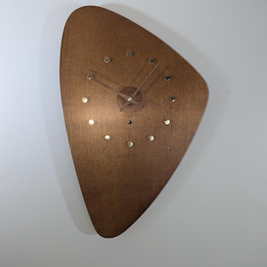 Könnte beinhalten: Eine braune, asymmetrische, dreieckige Wanduhr mit glatter Holzmaserung. Die Uhr verfügt über zwölf kleine, kreisförmige, metallfarbene Stundenmarkierungen und zwei dünne, passende metallfarbene Zeiger in der Mitte des Zifferblatts.