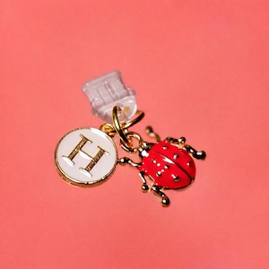 May include: A gold-toned charm with a red ladybug and a white circular pendant with the letter "H" in gold. The ladybug has black spots and gold legs. The charm is attached to a clear plastic clip, all against a pink background.
