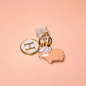 Pode incluir: Um berloque dourado com um círculo branco com a letra "H" e um berloque em forma de ovelha. A ovelha é cor de pêssego com uma cabeça branca e contorno dourado. Os berloques são conectados por um anel dourado.