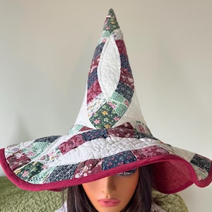 Pode incluir: Um chapéu de bruxa acolchoado feito à mão com uma aba larga. O chapéu apresenta um design de patchwork com padrões florais e geométricos em tons de bordô, verde e branco. A aba é forrada com um tecido bordô liso e o chapéu tem uma coroa alta e pontiaguda.