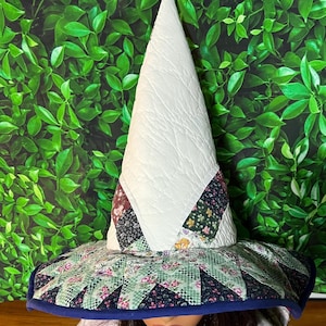 Pode incluir: Um chapéu de bruxa feito à mão com um cone acolchoado branco e uma aba larga. A aba apresenta um design de patchwork com padrões florais e geométricos em tons de verde, rosa e preto, delineados com uma borda azul. Um acessório único.