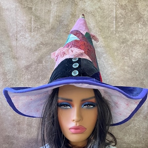 Puede incluir: Un sombrero de bruja caprichoso con ala ancha y corona alta y puntiaguda. El sombrero presenta un diseño de patchwork en tonos de rosa, morado y negro, con botones decorativos y ribete de encaje. El ala está forrada con una tela estampada y ribeteada en morado.