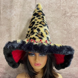 May include: A whimsical witch hat with a leopard print cone and a wide brim trimmed with black faux fur. The brim features red ear-shaped accents and is adorned with a delicate garland of red berries. A unique costume accessory.
