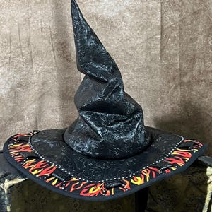 Puede incluir: Un sombrero puntiagudo negro con un patrón floral texturizado en relieve en la copa y el ala. El borde exterior del ala está decorado con un estampado de llamas vibrante en tonos rojos, naranjas y amarillos, acentuado por pequeñas pestañas de tela negra.