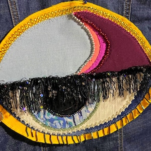 Può includere: Una toppa ricamata a forma di occhio, con bordo giallo, frange nere e un ciondolo pendente. L'occhio presenta sezioni azzurre, viola, rosa e bianche, con dettagli di perline e paillettes. La toppa è cucita su una giacca di jeans.
