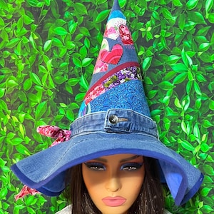 Pode incluir: Um chapéu de bruxa artesanal com aba de ganga e cone de retalhos. O cone apresenta detalhes florais e em forma de coração. Uma faixa de ganga com detalhe de botão circunda a base, e uma fita rosa adiciona um toque lúdico.