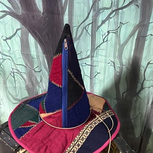 May include: A conical hat constructed from quilted fabric patches in shades of navy blue, deep red, black, and dark green. The hat features a prominent vertical blue zipper on the front and a wide brim edged in bright pink with decorative stitching.