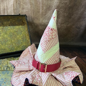 Könnte beinhalten: Ein skurriler Hexenhut mit rotem Gürtel und goldener Schnalle. Der Hut besteht aus gestepptem Stoff mit roten und weißen Blumen- und Streifenmustern. Der sternförmige Rand befindet sich auf einem Vintage-Koffer.