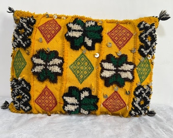 Può includere: Un cuscino rettangolare giallo con una superficie strutturata. Presenta motivi geometrici rossi, verdi e bianchi, con nappe bianche e nere agli angoli. Paillettes decorative sono sparse sulla superficie.