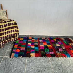 Peut inclure: Un tapis rectangulaire coloré avec un motif patchwork. Le tapis présente une variété de couleurs, dont le rouge, le bleu, le vert et le violet. Un cadre en bois avec de la laine est posé sur une table recouverte d'un tissu à carreaux jaune et marron.
