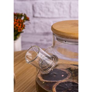 Könnte beinhalten: Eine transparente Glas-Teekanne mit Bambusdeckel, gefüllt mit Tee und getrockneten Zitrusfrüchten. Die Teekanne hat einen eingebauten Infuser und steht auf einer Holzoberfläche. Der Hintergrund zeigt eine weiße Ziegelmauer und eine kleine Pflanze.