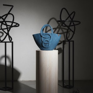 May include: A blue woven tote bag with unique handle detailing, displayed on a cylindrical stone pedestal. The bag's handles feature a looped design. Abstract black metal sculptures flank the pedestal, creating a modern aesthetic.