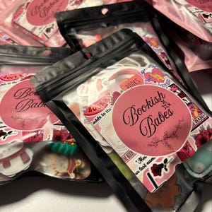 Può includere: Diverse buste di plastica trasparente piene di adesivi, con un design rosa e nero. Le buste hanno un'etichetta rosa con le parole "Bookish Babes" in corsivo. Gli adesivi hanno vari disegni, tra cui fiori e la parola "BOOKS".