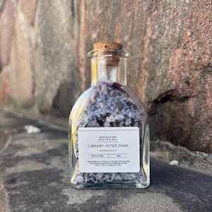 Könnte beinhalten: Eine klare Glasflasche mit Korken, gefüllt mit grauen und weißen Kristallen. Das Etikett lautet "LIBRARY AFTER DARK". Die Flasche steht auf einer grauen Oberfläche mit strukturiertem Steinhintergrund.