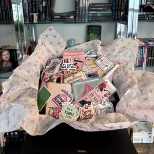 Puede incluir: Una caja de regalo llena de varios artículos, incluyendo pegatinas, marcadores y calcetines rosas. Las pegatinas tienen frases como "I'm a Kindle Girlie" y "Spicy Books". La caja está forrada con papel de seda decorado con pequeños lazos rojos.
