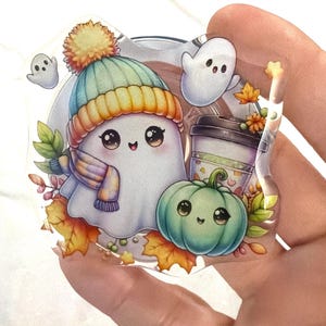 Puede incluir: Un dije de acrílico transparente con un fantasma de dibujos animados que lleva un gorro de punto azul y amarillo y una bufanda a cuadros, acompañado de una pequeña calabaza verde, una taza de café y dos pequeños fantasmas flotantes.