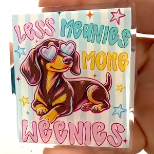 May include: A square acrylic keychain featuring a brown and black dachshund wearing pink heart-shaped sunglasses. The background has light blue stripes and colorful stars. The text reads Less Meanies More Weenies in pink and yellow lettering.