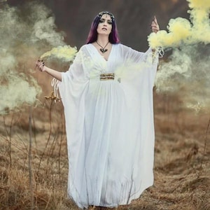 May include: A woman in a flowing white dress with a gold-toned belt and a V-neckline. She has purple hair and a headpiece. Yellow smoke billows around her, creating a mystical atmosphere. She is holding a smoke bomb in each hand.
